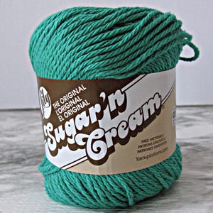May include: A ball of teal-colored Sugar'n Cream yarn. The label is brown and white, with the brand name and text that reads "The Original". The yarn is tightly wound and ready for crafting projects.