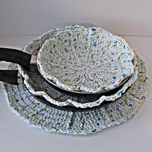 May include: A set of three crocheted pot holders in a speckled white, blue, and green pattern. The pot holders are circular with a scalloped edge, designed to fit around the base of a pan. The handles of the pans are black.