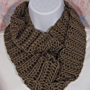 May include: A brown, handmade crochet infinity scarf. The scarf is displayed on a white mannequin head. The crochet stitch creates a textured, geometric pattern. The scarf is a warm, neutral color, suitable for fall or winter.