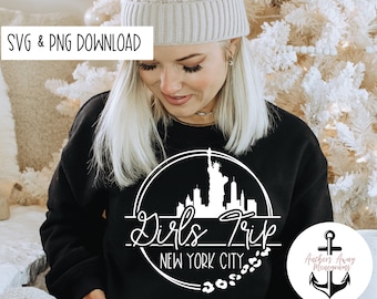 May include: A black sweatshirt with a white graphic design of the New York City skyline, including the Statue of Liberty, with the text "Girls Trip New York City" in a circle. The graphic also includes a line of white paw prints. The sweatshirt is being worn by a person with blonde hair and a white beanie.