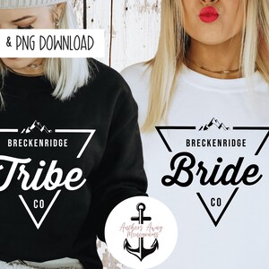 May include: Two women wearing black and white sweatshirts with a mountain range graphic and the text "Breckenridge" and "Tribe" or "Bride" in a white triangle. The text "CO" is below the triangle. The women are standing in front of a wood panel background.