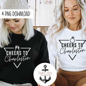 May include: Two white sweatshirts with black text. The text on one sweatshirt says "Cheers to Charleston" with a ring and a triangle. The text on the other sweatshirt says "Cheers to Charleston" with two champagne glasses and a triangle.