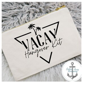 Op de afbeelding: Witte canvas toilettas met een zwart grafisch ontwerp van palmbomen en de tekst "Vacay Hangover Kit" in een driehoek. De tas heeft een gouden rits.