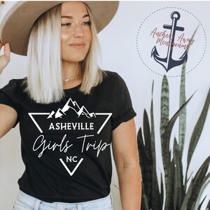 May include: Black t-shirt with a white graphic design of mountains and a triangle with the text "Asheville Girls Trip NC".