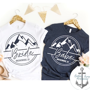 May include: Two white t-shirts with a navy blue mountain range design. The left shirt says "Bride" and the right shirt says "Babe". Both shirts have the text "Breckenridge, CO" underneath the design.