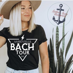 May include: A black t-shirt with a white graphic design that says "Asheville Bach Tour NC" inside a triangle with a lightning bolt above it.