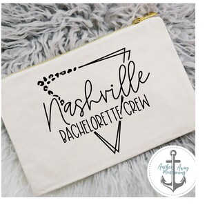 May include: White canvas makeup bag with a black graphic design that says "Nashville Bachelorette Crew" with a geometric design and leopard print accents. The bag has a gold zipper.