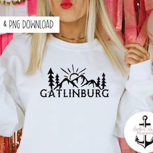 Può includere: Felpa bianca con un design grafico nero di una catena montuosa con un sole e alberi. Il testo "Gatlinburg" è sotto la grafica. La grafica è centrata sulla felpa. La felpa è indossata da una persona.