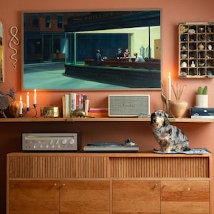 May include: A living room scene with a wooden cabinet, a painting of a diner, and a dachshund dog. The cabinet has a record player and a speaker on top. The painting is above a shelf with books and candles.