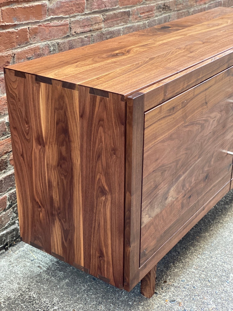K&ouml;nnte beinhalten: Ein dunkelbraunes Holzkabinett mit schlichtem Design und glatter Oberfl&auml;che. Das Kabinett hat zwei T&uuml;ren und steht auf vier kurzen Beinen.