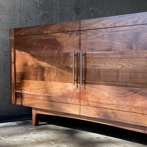 K&ouml;nnte beinhalten: Ein Holzschr&auml;nk mit zwei T&uuml;ren und silbernen Griffen. Der Schrank ist aus dunklem Holz mit nat&uuml;rlicher Maserung. Der Schrank steht auf einem Betonboden.