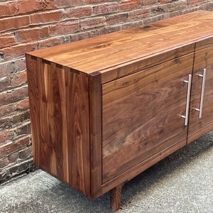 K&ouml;nnte beinhalten: Ein dunkelbraunes Holzschr&auml;nkchen mit zwei T&uuml;ren und silbernen Griffen. Das Schr&auml;nkchen hat ein schlichtes Design und ist aus massivem Holz gefertigt. Das Schr&auml;nkchen steht auf einem Betonboden.