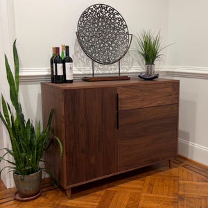 May include: A dark wood cabinet with a drawer and door, topped with a decorative metal sculpture, wine bottles, and a potted plant. The cabinet sits on a parquet floor. A tall potted snake plant is to the left.