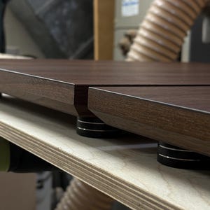 May include: Close-up of a dark brown wooden table top with a beveled edge. The table is supported by black and silver cylindrical feet. The wood grain is visible, and the table appears to be new.