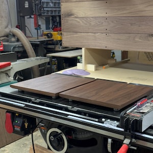 May include: Two square dark brown walnut wood panels with smooth surfaces and rounded edges are positioned on a black metal table saw surface. The panels feature a visible wood grain pattern and are elevated slightly on small circular supports.