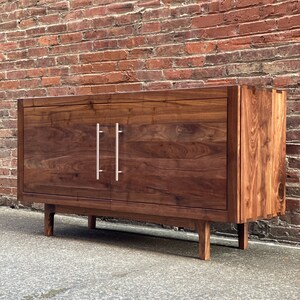 K&ouml;nnte beinhalten: Ein dunkelbraunes Holzschr&auml;nkchen mit zwei T&uuml;ren und silbernen Griffen. Das Schr&auml;nkchen hat ein schlichtes Design mit einer rechteckigen Form und vier Beinen. Das Schr&auml;nkchen steht auf einem Betonboden vor einer Backsteinwand.
