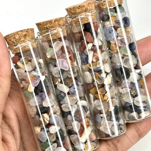 Puede incluir: Cuatro pequeñas botellas de vidrio con tapas de corcho llenas de piedras pulidas de colores. Las botellas están dispuestas en fila, con la botella más grande en la parte posterior y la más pequeña en la parte delantera.