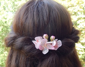 May include: A pink flower hair clip with three blossoms and two buds. The clip is in a woman's brown hair.