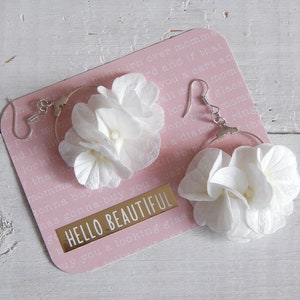 May include: A pair of hoop earrings with white flower accents. The earrings are on a pink card with the text "HELLO BEAUTIFUL" printed on it.