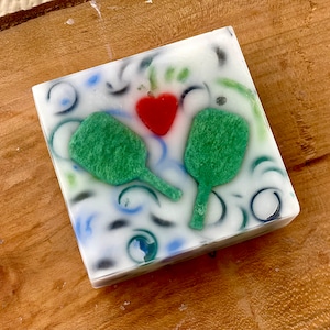 May include: A white square soap bar with a red heart and two green paddle shapes embedded in the surface. The soap has a swirling pattern of blue, green, and black.