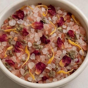 Pode incluir: Uma tigela de cerâmica branca cheia de cristais de sal rosa e branco, pétalas de rosa vermelhas secas, pétalas de flores amarelas e ervas verdes. O produto de banho é uma foto em close-up.