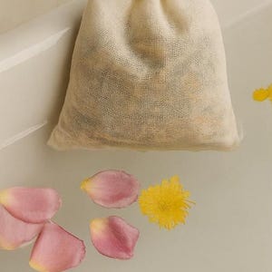 Pode incluir: Um saco de banho de musselina bege cheio de ervas secas flutua na água, rodeado por pétalas de flores rosa e amarelas. O saco é amarrado no topo, sugerindo um produto de spa ou autocuidado.