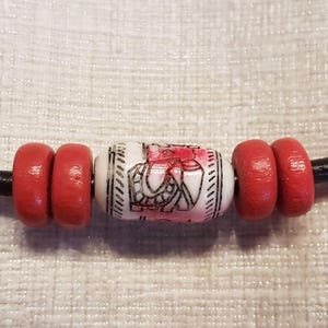 Könnte beinhalten: Eine Halskette aus schwarzem Lederband mit einer weißen Perle mit roten Akzenten und zwei roten Perlen auf jeder Seite.