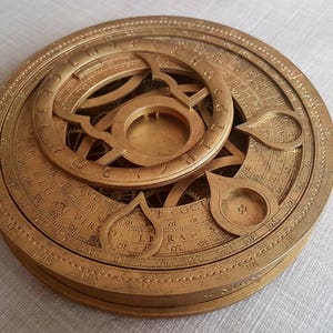 Könnte beinhalten: Ein goldfarbener, komplizierter, runder Astrolab mit einem drehbaren Zifferblatt und gravierten Markierungen. Das Astrolab ist ein historisches Navigationsinstrument, das zur Bestimmung der Position von Himmelskörpern verwendet wurde.