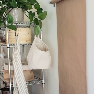 Op de afbeelding: Een metalen rek met een hangende canvas tas, een gestreepte schort en een houten doos. Een rol bruin papier is aan de muur boven het rek bevestigd.