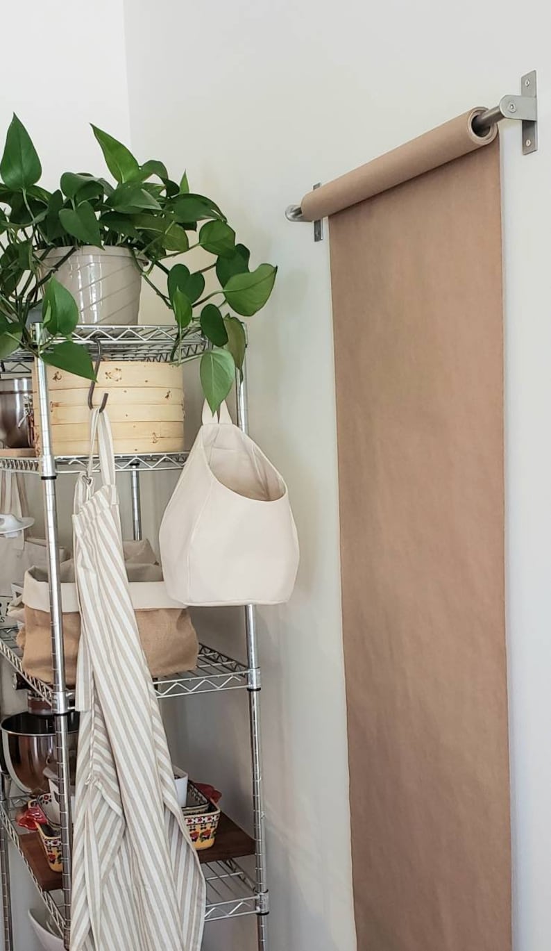 Op de afbeelding: Een metalen rek met een hangende canvas tas, een gestreepte schort en een houten doos. Een rol bruin papier is aan de muur boven het rek bevestigd.