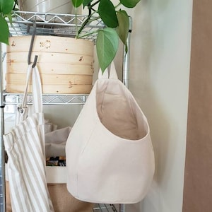 Op de afbeelding: Een witte hangende opbergmand van stof met een brede opening. De mand hangt aan een metalen rek. Op de achtergrond staat een plant in een pot met groene bladeren.