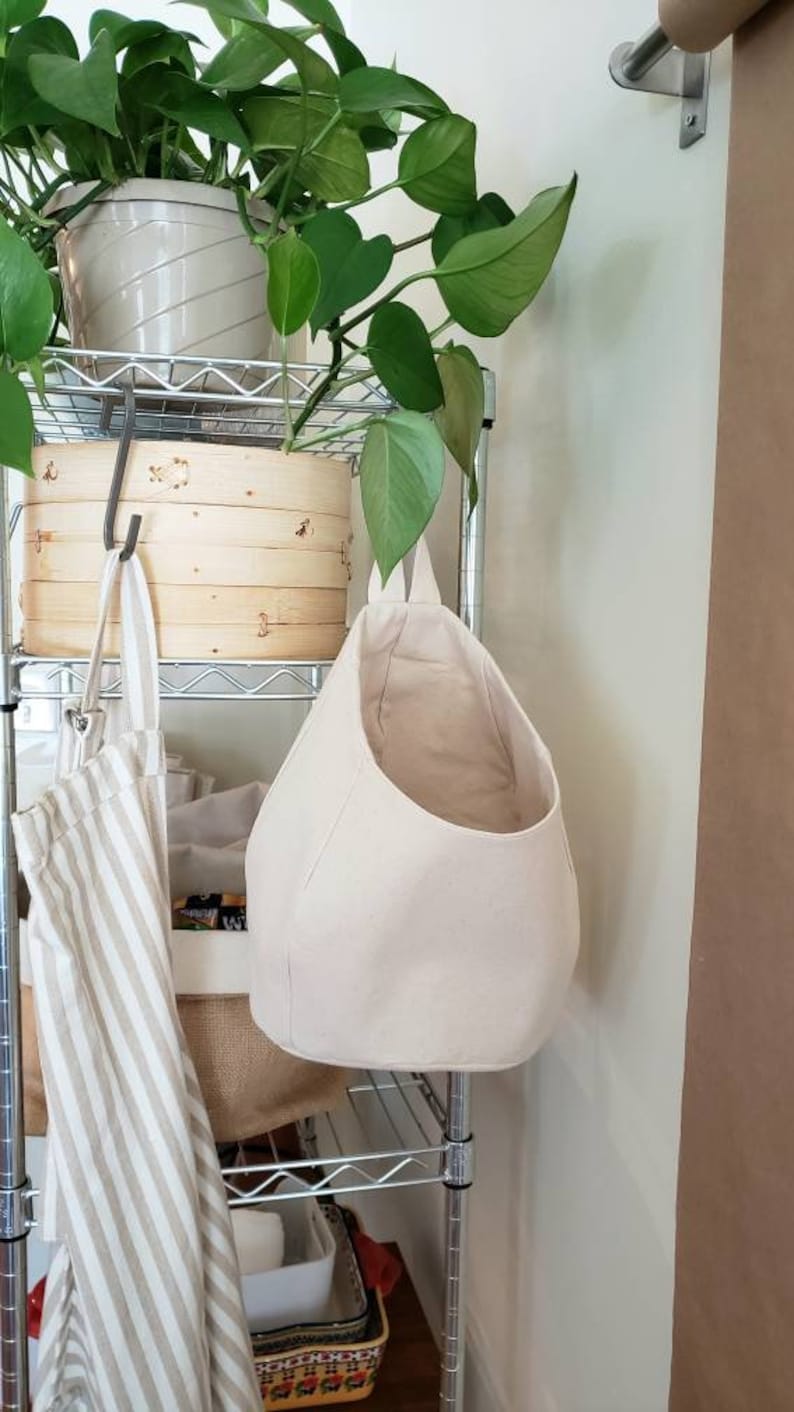 Op de afbeelding: Een witte hangende opbergmand van stof met een brede opening. De mand hangt aan een metalen rek. Op de achtergrond staat een plant in een pot met groene bladeren.