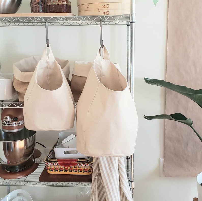 Op de afbeelding: Twee cr&egrave;mekleurige canvas opbergtassen hangen aan een metalen plank. De tassen hebben een afgeronde bodem en zijn gemaakt van een zachte, natuurlijke stof. De tassen hangen aan haken die aan de plank zijn bevestigd.