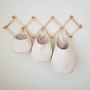 Op de afbeelding: Drie cr&egrave;mekleurige canvas opbergmanden hangen aan een houten wandorganizer. De organizer heeft een ruitpatroon en is gemaakt van natuurlijk hout.