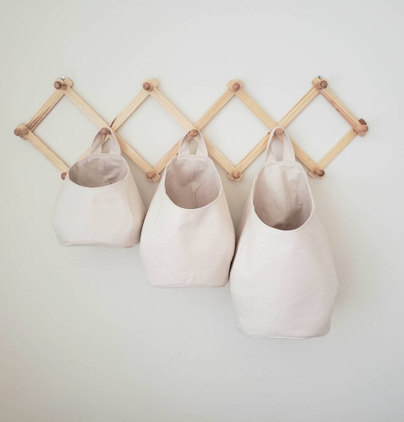 Op de afbeelding: Drie cr&egrave;mekleurige canvas opbergmanden hangen aan een houten wandorganizer. De organizer heeft een ruitpatroon en is gemaakt van natuurlijk hout.