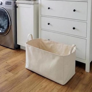 May include: A rectangular off-white canvas laundry basket with two fabric handles on the sides. The basket has a structured base and soft, collapsible walls, providing a simple storage solution for household items or clothing in a room.