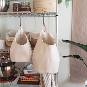 Op de afbeelding: Twee beige canvas hangende opbergtassen met een metalen haak aan een metalen rek. De tassen hangen aan de haken en zijn leeg. De planken zijn gevuld met verschillende items, waaronder een houten kom, potten en een keukenmixer.