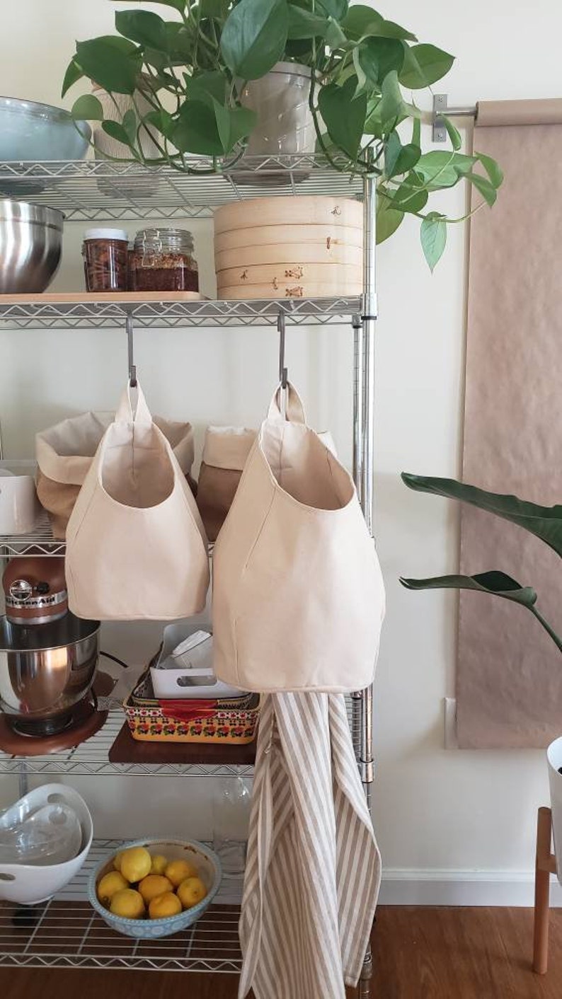 Op de afbeelding: Twee beige canvas hangende opbergtassen met een metalen haak aan een metalen rek. De tassen hangen aan de haken en zijn leeg. De planken zijn gevuld met verschillende items, waaronder een houten kom, potten en een keukenmixer.