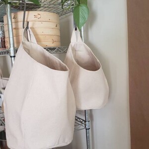 Op de afbeelding: Twee beige canvas hangende opbergmanden met een metalen plank erachter. Een plant in een pot met groene bladeren hangt boven de plank.