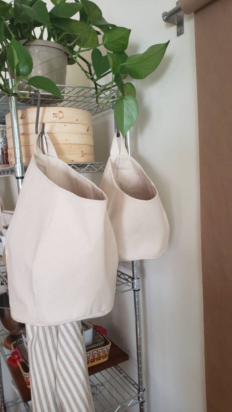 Op de afbeelding: Twee beige canvas hangende opbergmanden met een metalen plank erachter. Een plant in een pot met groene bladeren hangt boven de plank.