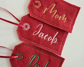 May include: Three red felt gift tags with embroidered names: "Mom" in gold, "Jacob" in white, and "Dad" in green. Each tag has a decorative stitched border and a red ribbon for hanging.