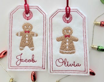 May include: Two white felt gift tags with red stitched borders and gingerbread man embroidery. The tags have red ribbon loops and the names "Jacob" and "Olivia" embroidered in red. Christmas lights are in the background.
