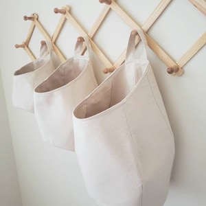 Op de afbeelding: Drie off-white canvas opbergmanden die aan een houten wandrek hangen. De manden hebben een ronde bodem en een enkele lusvormige handgreep.