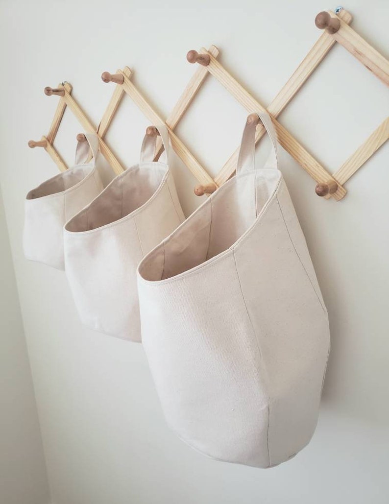 Op de afbeelding: Drie off-white canvas opbergmanden die aan een houten wandrek hangen. De manden hebben een ronde bodem en een enkele lusvormige handgreep.