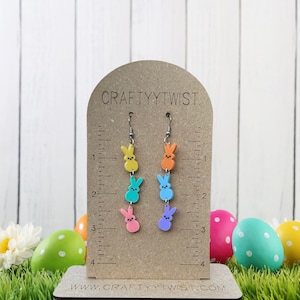 May include: A pair of dangle earrings featuring four pastel-colored bunny charms. The charms are yellow, blue, pink, and orange. The earrings are displayed on a brown card with the text "CRAFTYTWIST" and "WWW.CRAFTYTWIST.COM".