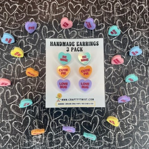 May include: A set of six pairs of handmade heart-shaped earrings. The earrings are made of plastic and have a variety of colors and sayings, including "Be Mine", "Cutie Pie", and "Love Bug".