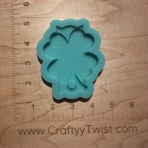 May include: A teal blue silicone mold shaped like a four-leaf clover with a small circle in the center. The mold is approximately 2 inches in diameter.
