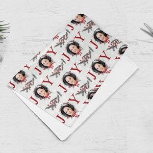 May include: White wrapping paper with a repeating pattern of a person's face in a wreath with red berries and green pine branches. The word "JOY" is repeated in red letters throughout the pattern.