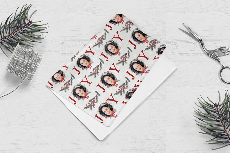 May include: White wrapping paper with a repeating pattern of a person's face in a wreath with red berries and green pine branches. The word "JOY" is repeated in red letters throughout the pattern.