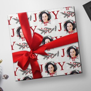 May include: White gift wrap with a red ribbon and a repeating pattern of the word "JOY" in red letters, a wreath with berries and pine branches, and a photo of a person in the center of each wreath.